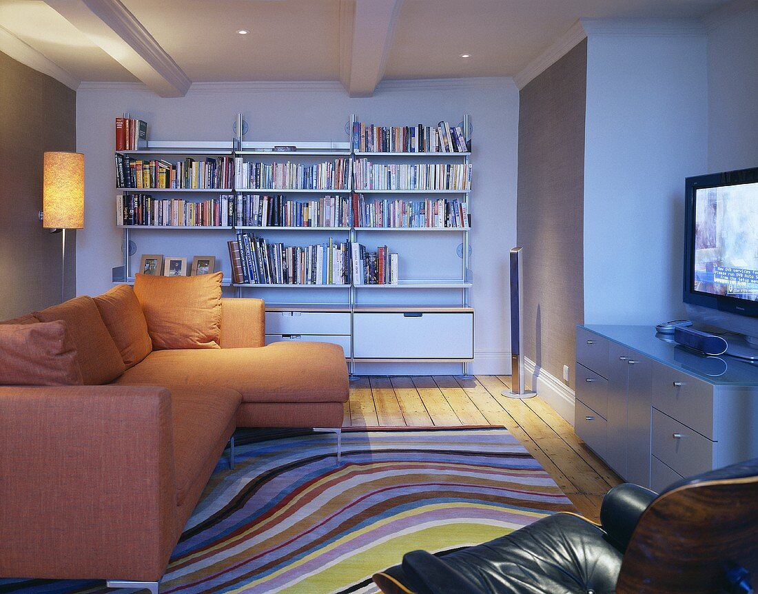 An open-plan living room with an orange corner sofa in front of a bookshelf and a rug with curved stripes