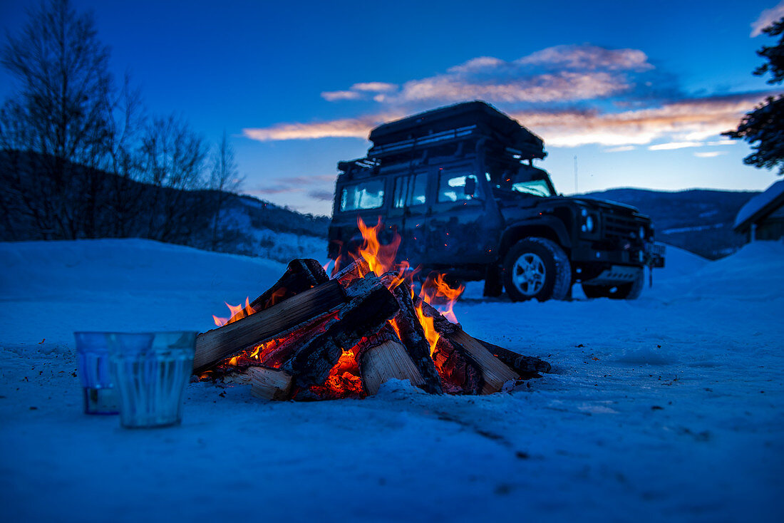 Lagerfeuer in Winterlandschaft mit Geländewagen im Hintergrund, Heggenes, Norwegen