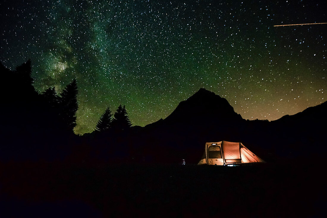 Illuminated tent stands in the night under the starry sky in front of a mountain silhouette. A shooting star flies into the picture. Obersee. Switzerland;