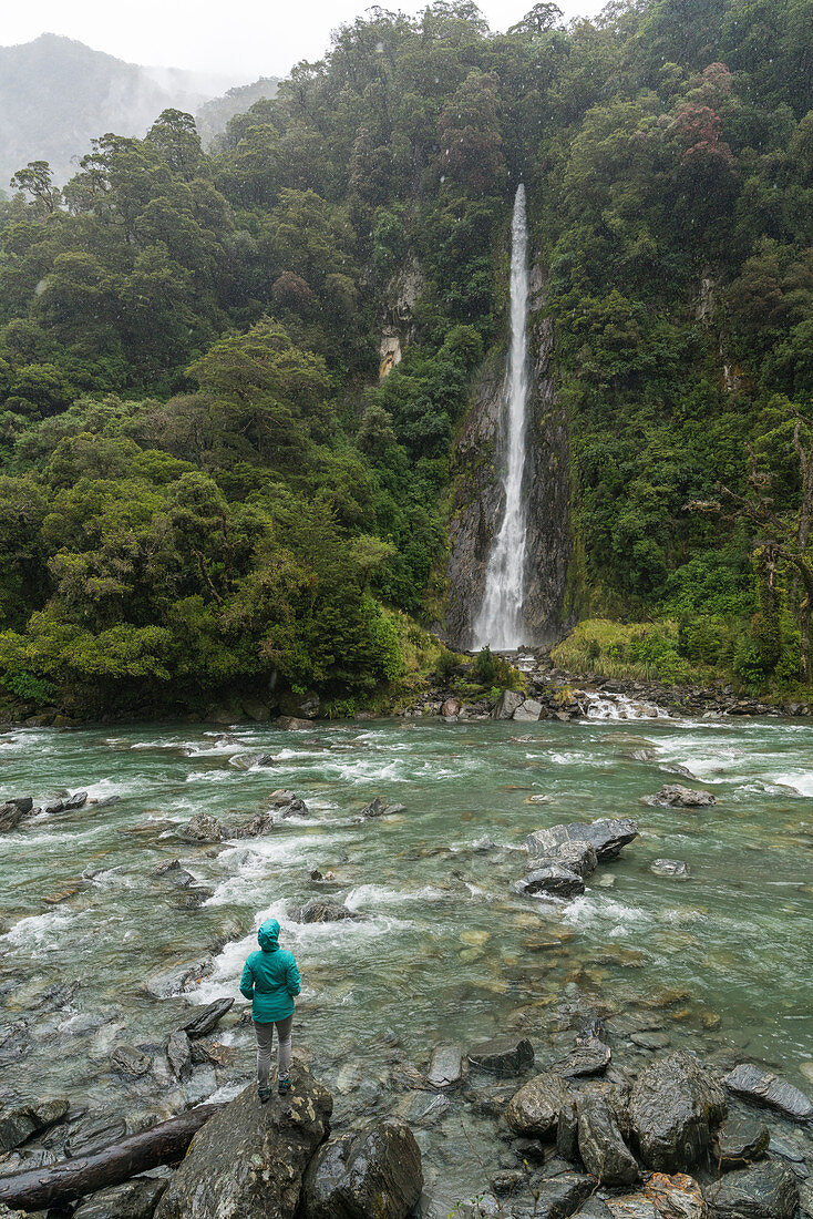 Tourist under the rain admiring Thunder Creek Falls. Mount Aspiring National Park, West Coast region, South Island, New Zealand.
