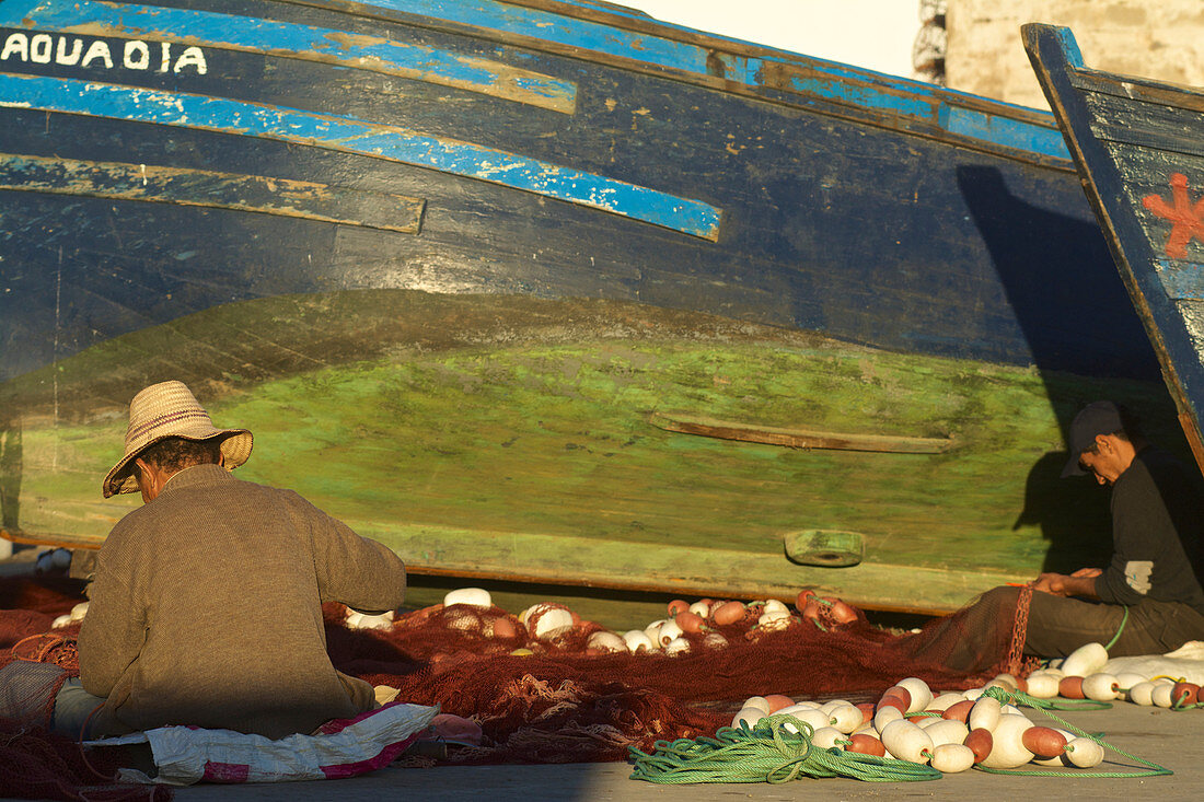 Fischer sitzen im Hafen von Essaouira auf dem Boden und reparieren Netze vor einem farbigen Boot, Essaouira, Marokko