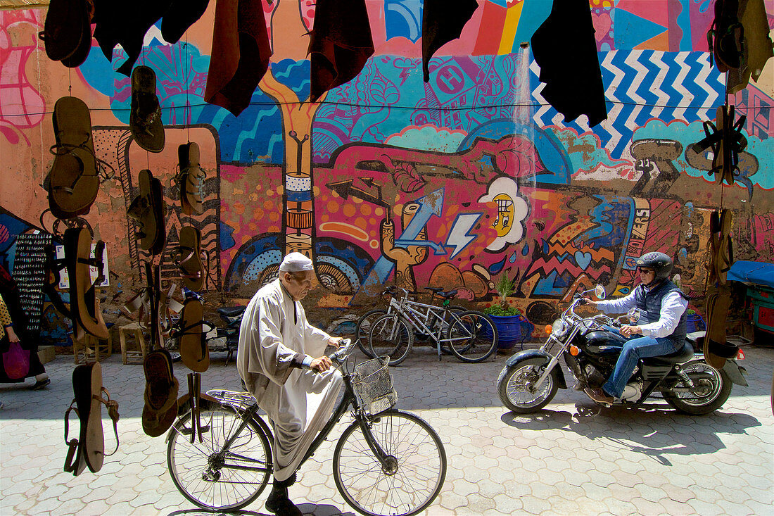 Alter Mann auf Fahrrad begegnet Tourist auf Motorrad vor einer bunt bemalten Wand in der Altstadt von Marrakesch, Marokko