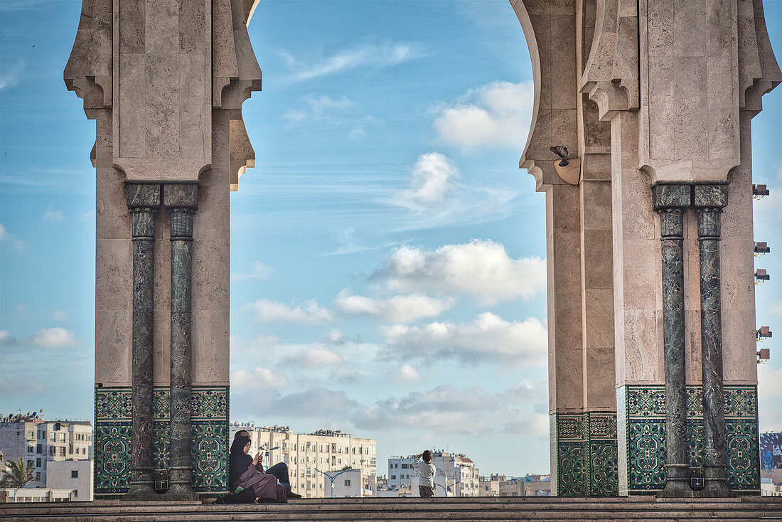 View through arches of the Hassan II mosque on modern buildings, woman in traditional clothing, Casablanca, Morocco