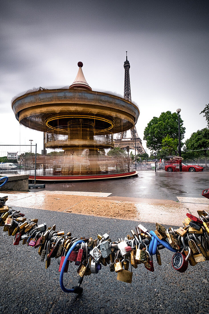 Carousel in front of the Eiffel Tower in rainy weather; in the foreground, love-locks are fastened to the chain of street-bluffing; Paris; Île-de-france; France