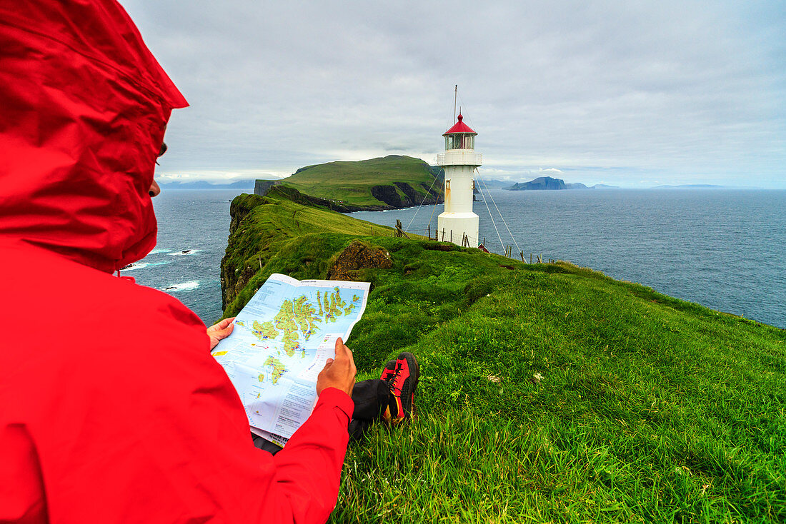 Hiker on cliffs looks at the map next to lighthouse, Mykines island, Faroe Islands, Denmark, Europe