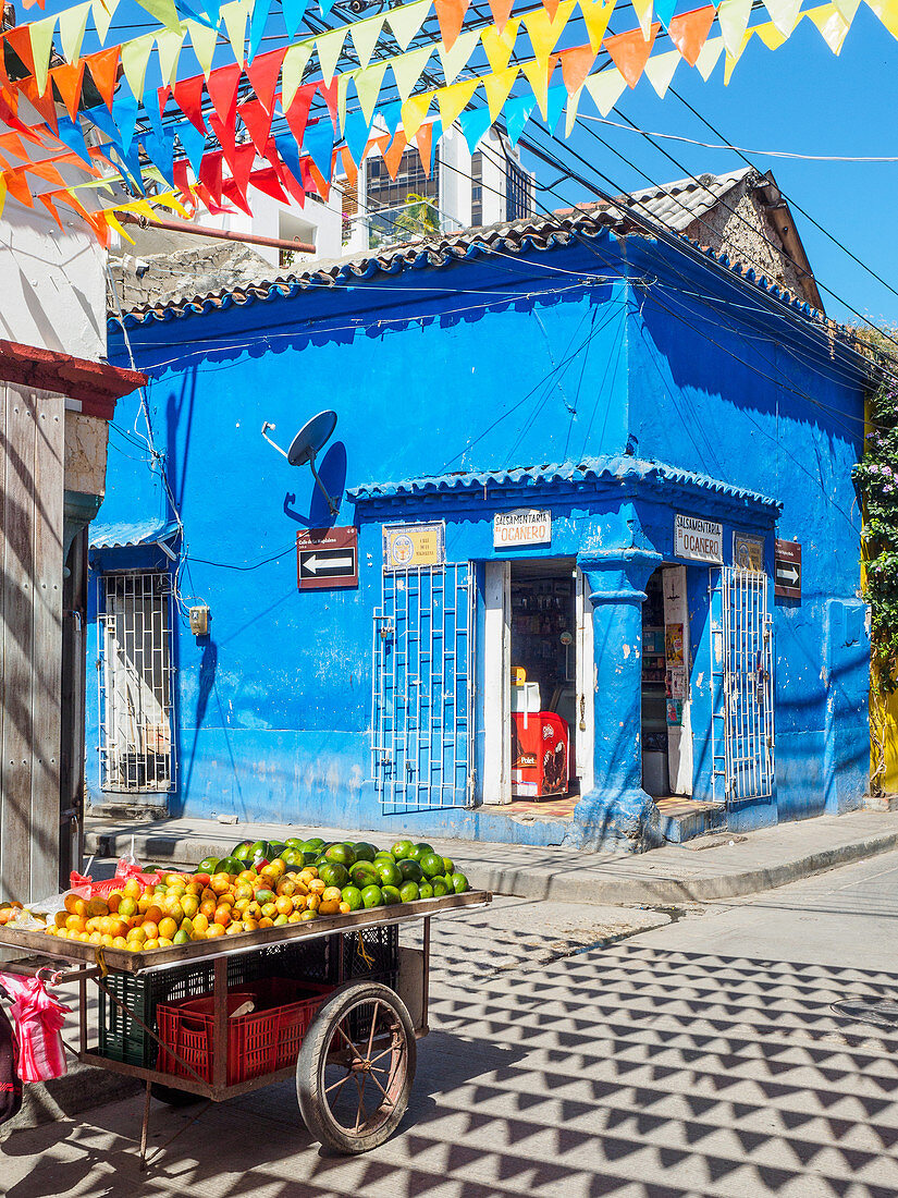 Bunte Fahnen, blaues Gebäude und Obstwagen an einer Straßenecke im Getsemani-Barrio, Cartagena, Kolumbien, Südamerika