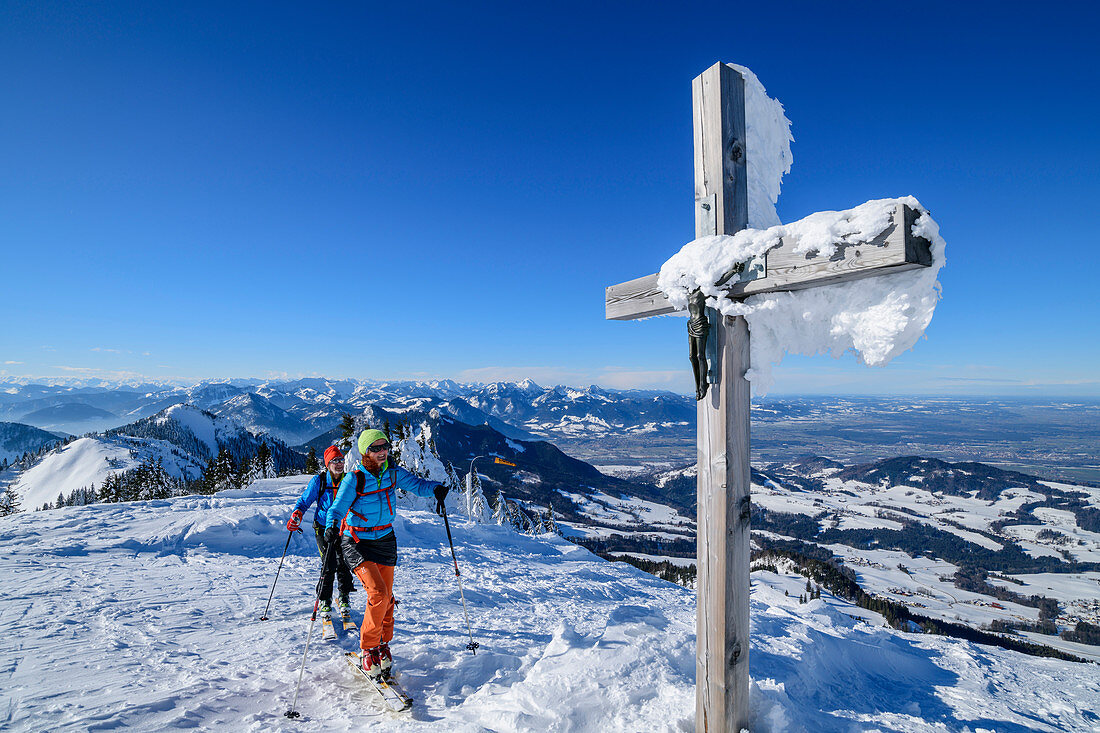 Mann und Frau auf Skitour gehen an verschneitem Gipfelkreuz vorbei, Hochries, Chiemgauer Alpen, Bayern, Deutschland