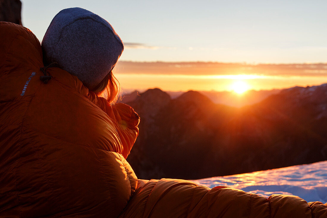 Woman in down sleeping bag looks at the rising morning sun, Gramai, Karwendel, Tyrol, Austria