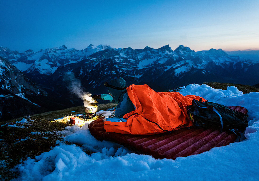 Young woman lies in her bivouac sack on a sleeping pad in the snow and boils water with a gas cooker in the glow of her headlamp in front of the Karwendel backdrop, Tyrol, Austria