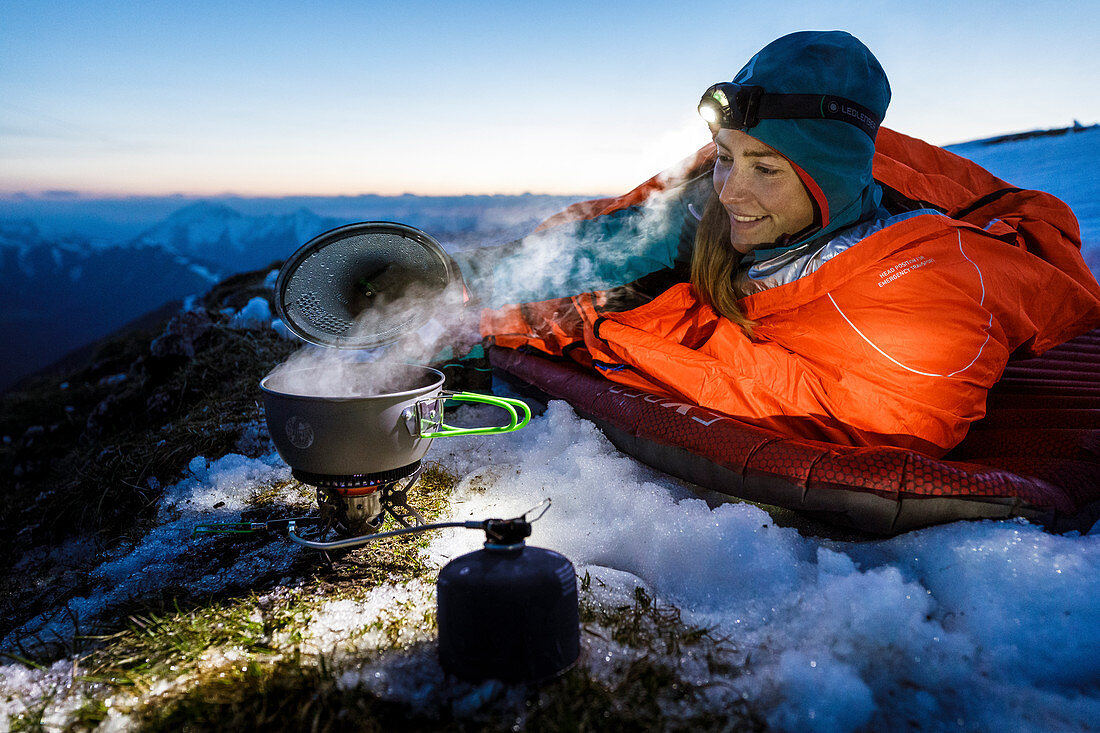 Young woman bivouacs on the summit on sleeping pad and in a bivouac sack and boils water with a gas stove, Karwendel, Tyrol, Austria
