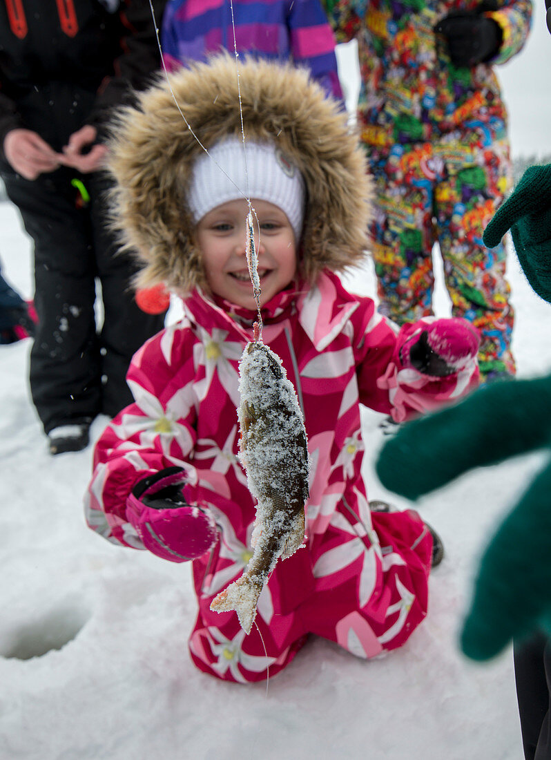 Kleinkind im Schneeanzug bewundert geangelten Fisch, Eisfischen in Finnland