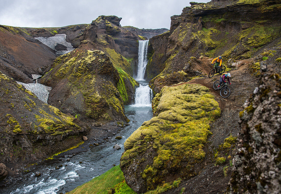 Zwei Mountainbiker fahren einen schmalen Pfad neben einem Wasserfall herunter, Landschaft in Island