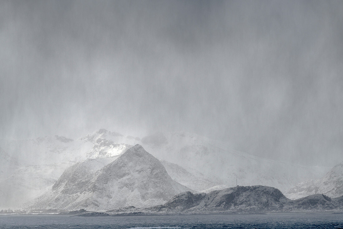 Schneesturm über dem Meer mit verschneiten Bergen im Hintergrund, Lofoten, Nordland, Norwegen