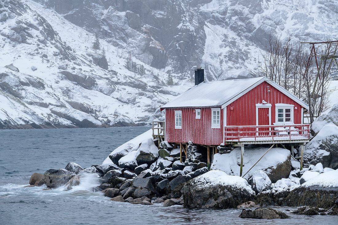 Norwegisches rotes Fischerhaus am Meer mit verschneiten Bergen im Hintergrund, Nusfjord, Lofoten, Nordland, Norwegen