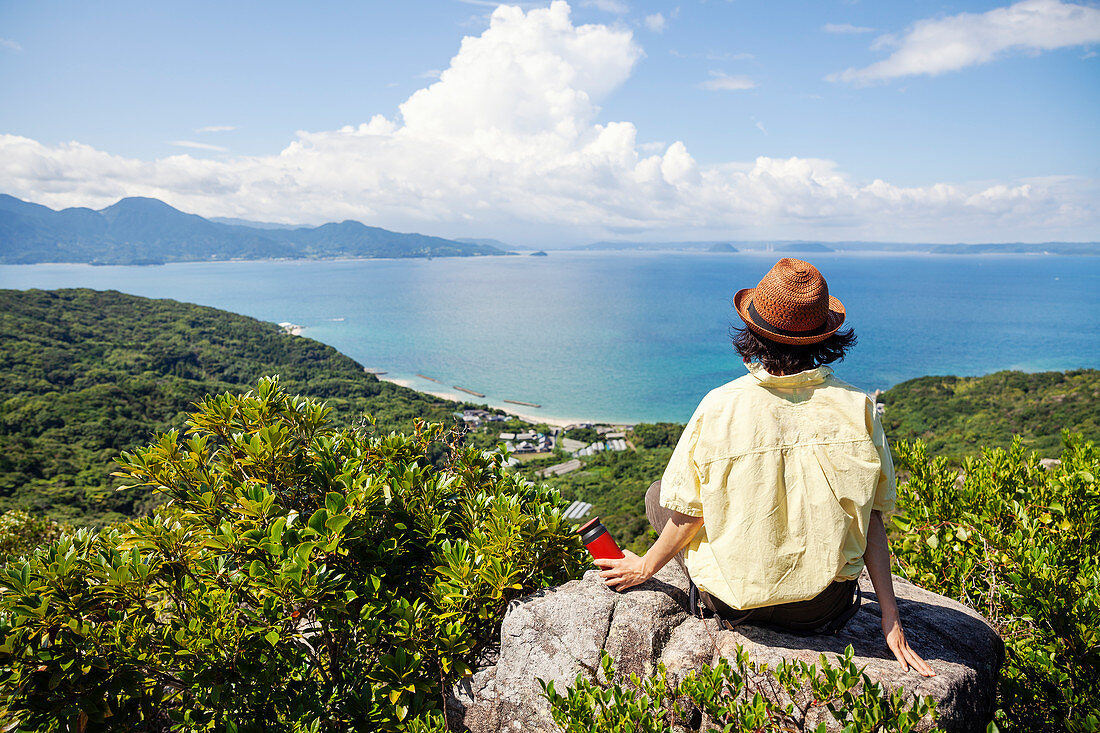 Japanische Frau mit Hut sitzt auf Felsen auf einer Klippe, Ozean im Hintergrund