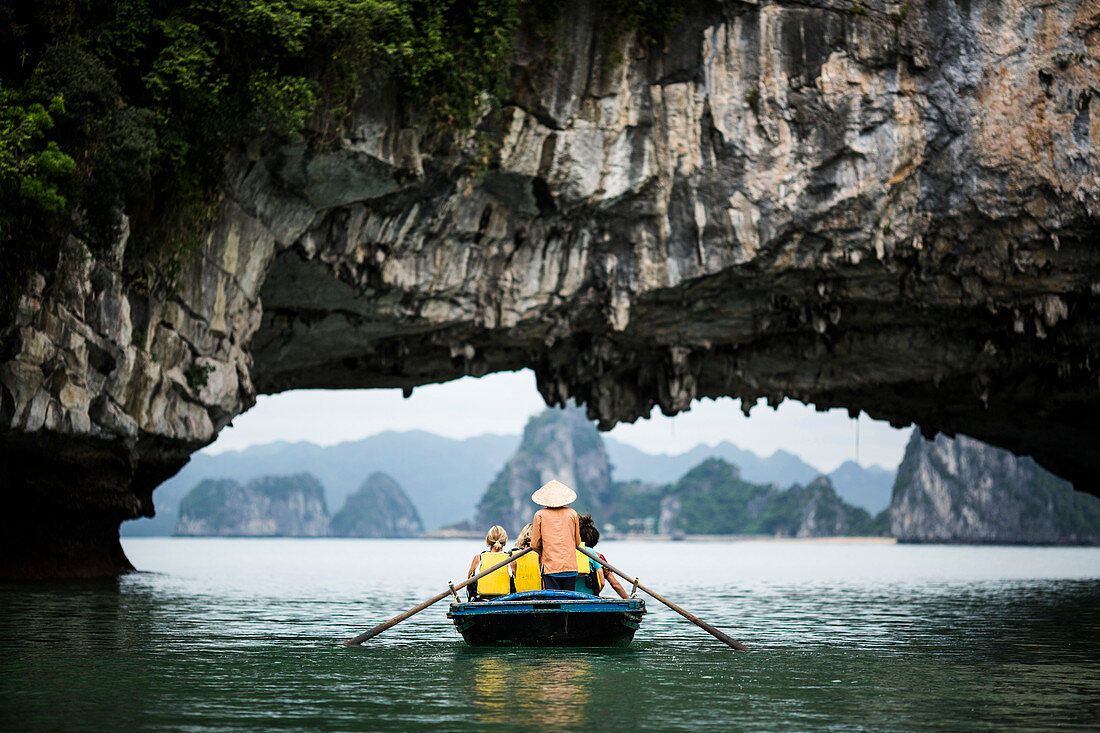 Rear view of man wearing straw hat transporting small group of people on a boat, rowing underneath natural rock arch.