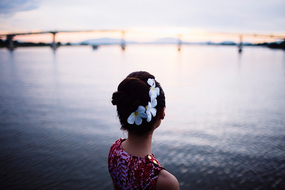 Rear view of woman with flowers in her hair, standing by the sea at sunset, bridge in the distance.