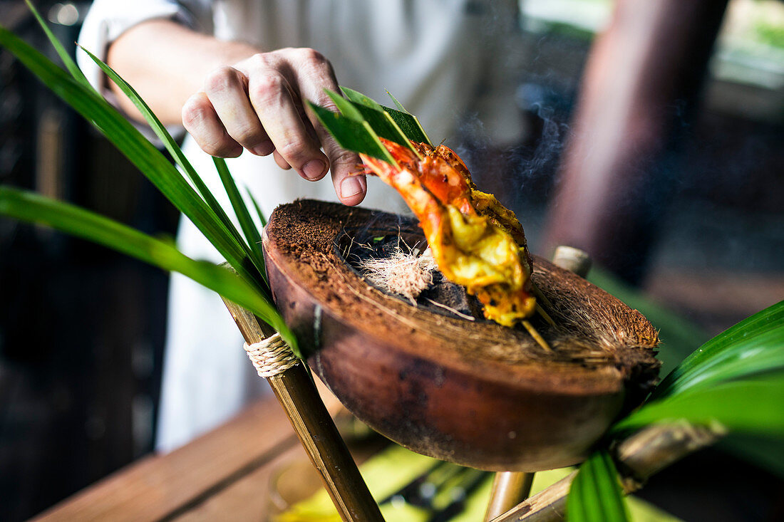 Close up of chef preparing charcoal-grilled prawn satay on a coconut shell.