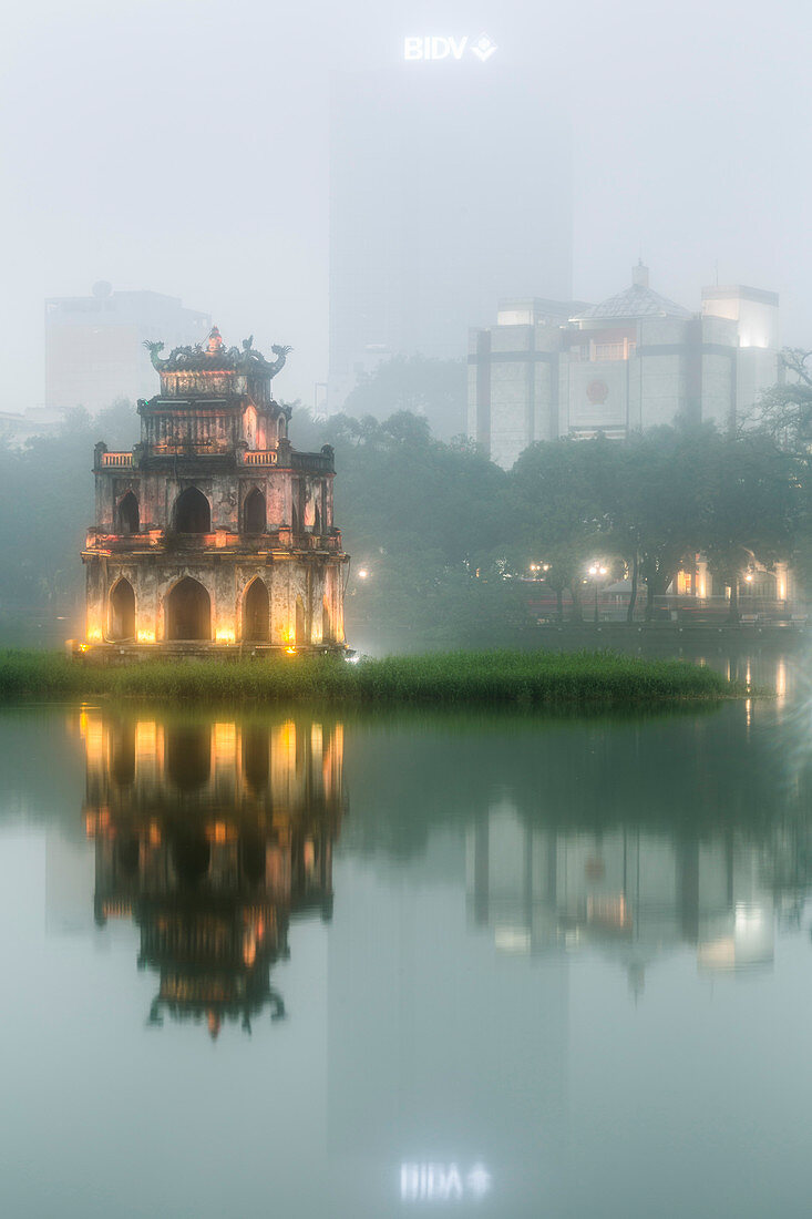 Exterior of illuminated temple reflected in a lake at dusk, skyscraper in the distance.