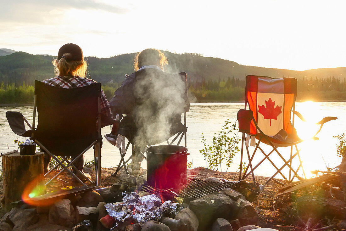 Two girls sit by the fire in the sunset and enjoy the view of the Yukon River. Canada, Whitehorse, Yukon