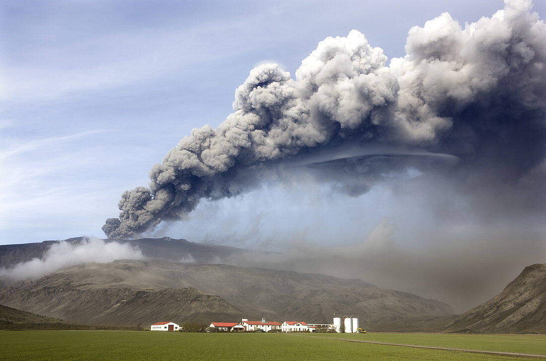 Wirtschaftsgebäude und grüne Felder mit Aschewolke des Eyjafjallajokull-Ausbruchs in der Ferne, bei Hella, südliches Gebiet, Island, Polarregionen
