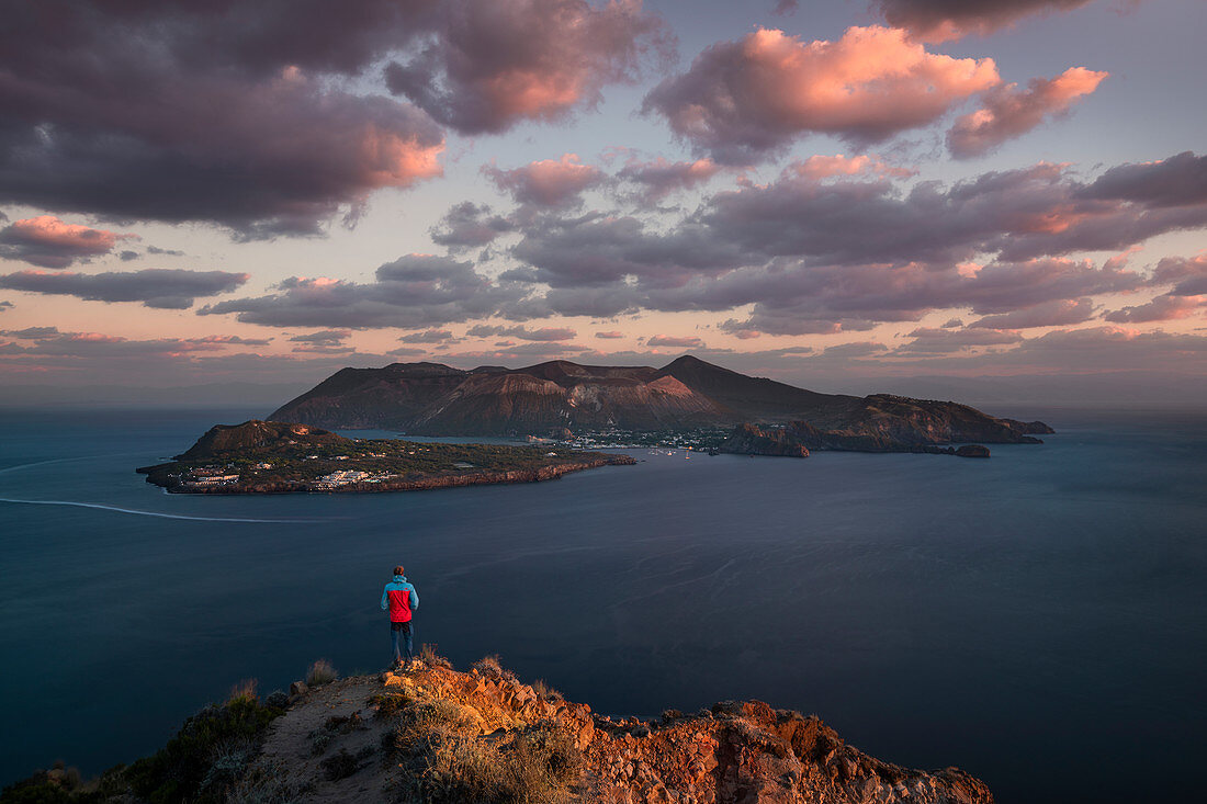 Mann mit roter Jacke an Küste von Lipari mit Blick auf Vulkaninsel Vulcano im Sonnenuntergang, Sizilien Italien\n