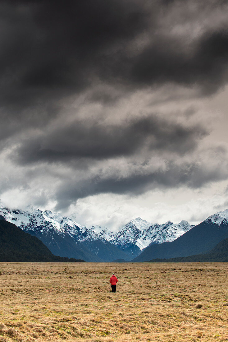 Man in red coat stood looking at snow capped mountains, Fiordland National Park, UNESCO World Heritage Site, South Island, New Zealand, Pacific
