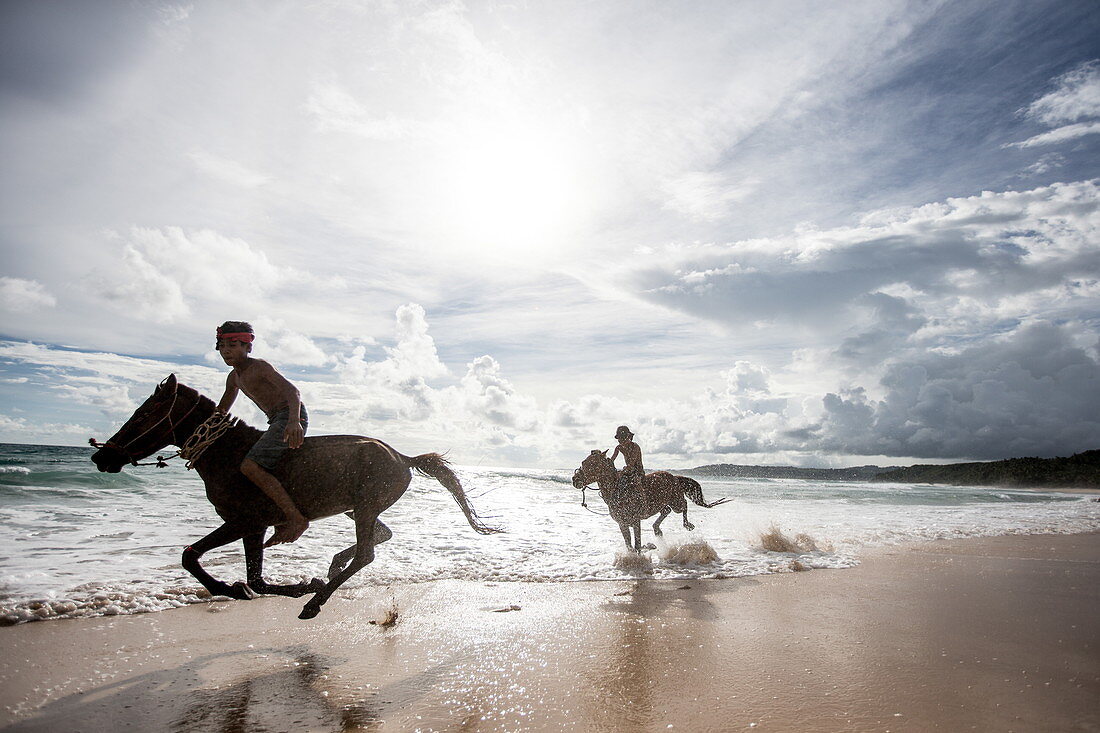 Zwei Jungen auf Pferden spielen im Ozean in Nihiwatu, Sumba, Indonesien, Südostasien, Asien