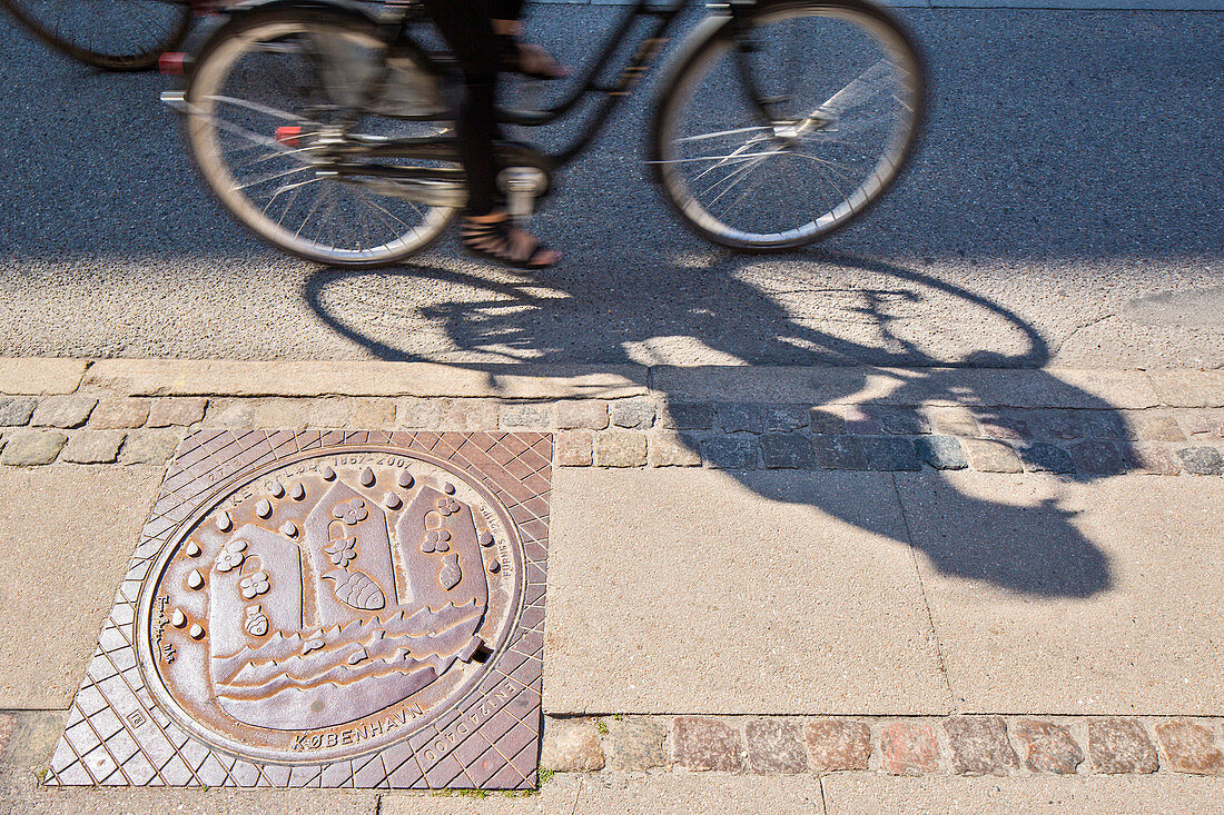 Kanalluke mit Kopenhagens Wappen, Pflaster, Schatten eines Bikers. Bredgade Straße, Kopenhagen, Seeland, Dänemark