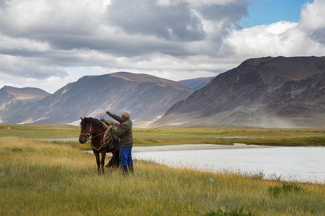 Reiter bereitet sein Pferd in der Nähe eines von Bergen umgebenen Sees, Tavan Bogd Massiv, Altai, Provinz Bayan-Olgii, Mongolei