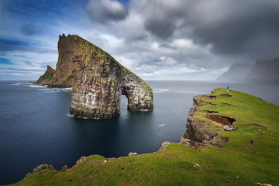 HIKER STANDING ON THE EDGE OF A GRASS-COVERED CLIFF CONTEMPLATING THE ARCHED SEA STACK OF DRANGARNIR, VAGAR, FAROE ISLANDS, DENMARK