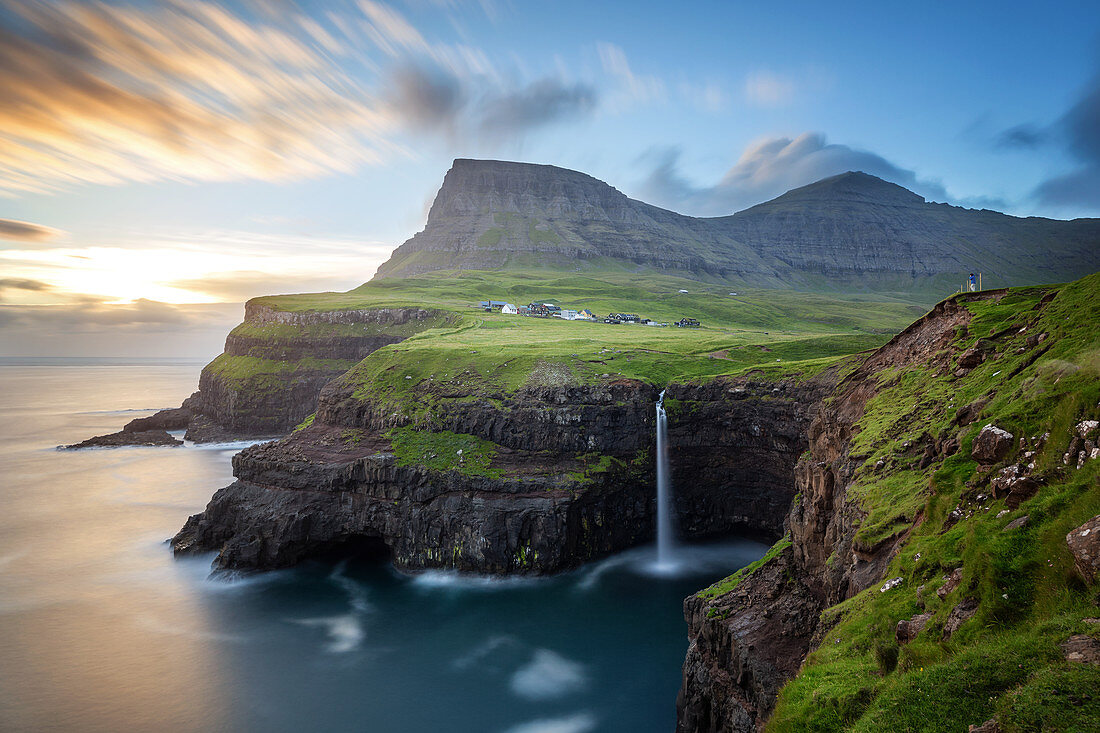 HIKER CONTEMPLATING THE MOUNTAINS, THE CLIFFS AND THE WATERFALL CASCADING INTO THE SEA BY THE VILLAGE OF GASADALUR, VAGAR, FAROE ISLANDS, DENMARK