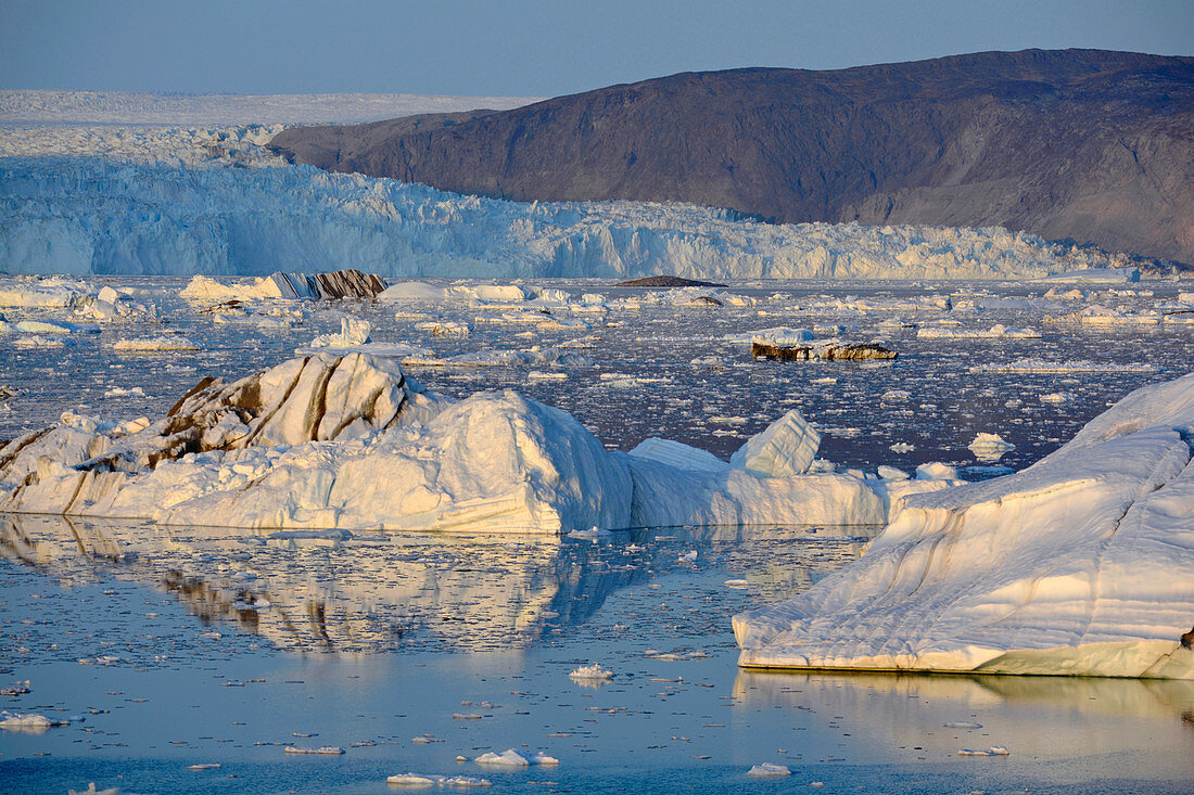 Eqi Glacier in West Greenland; Foothills of the inland ice in the background; floating icebergs and ice floes; Ridges with dark rocks; absolutely clear sky; the silhouettes of the ice mountains are reflected on the calm, blue water surface; Evening sun turns the ice surfaces light yellow