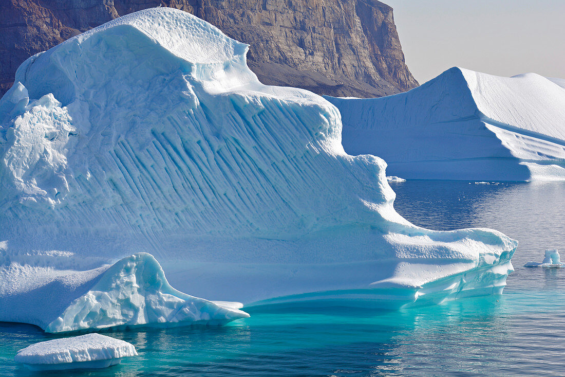 Icebergs off Uummannaq in West Greenland; diverse surfaces and structures; Ice under water glows intensely turquoise; steep, rocky mountain wall in the background; cloudless sky and absolutely calm water surface