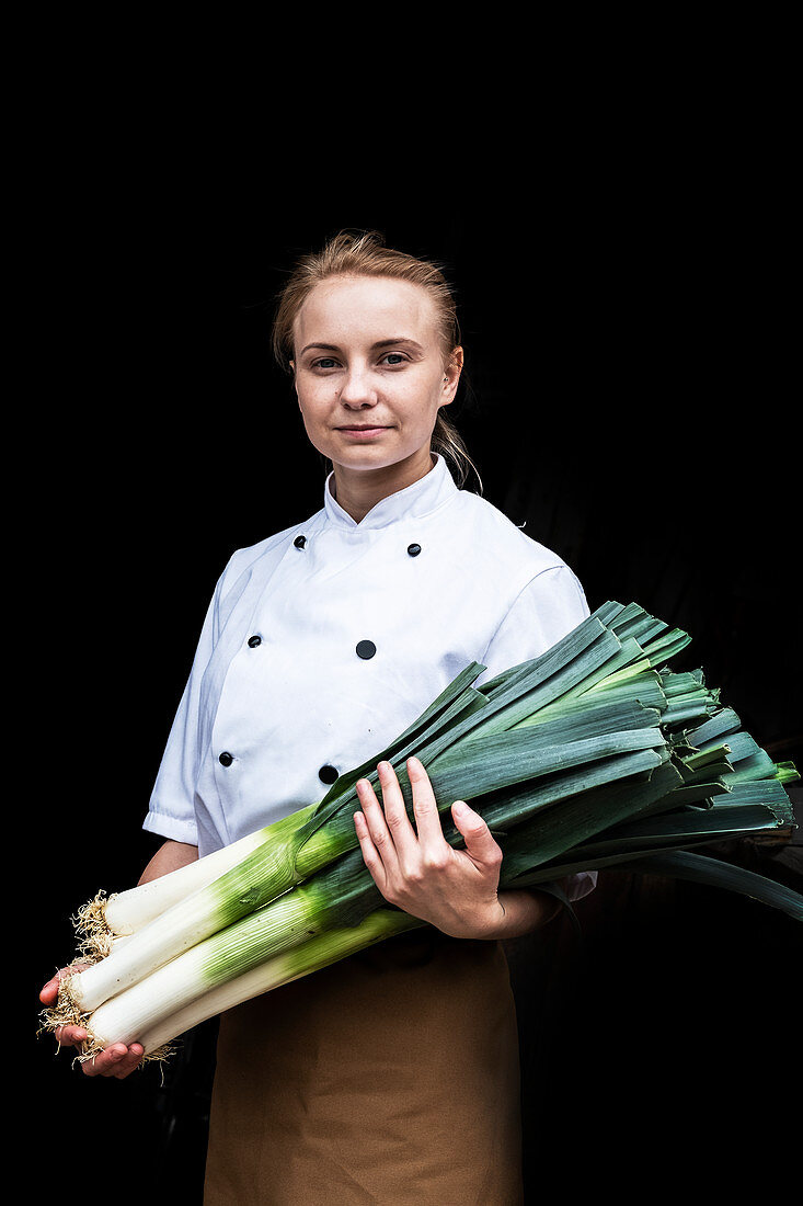 Woman wearing chef's jacket standing indoors, holding bunch of leeks, looking at camera.