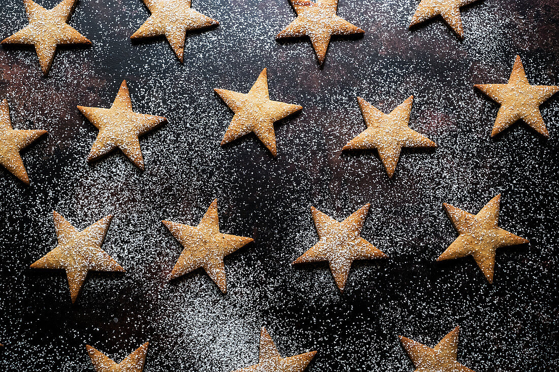 High angle close up of freshly baked star-shaped cookies dusted with icing sugar on black background.