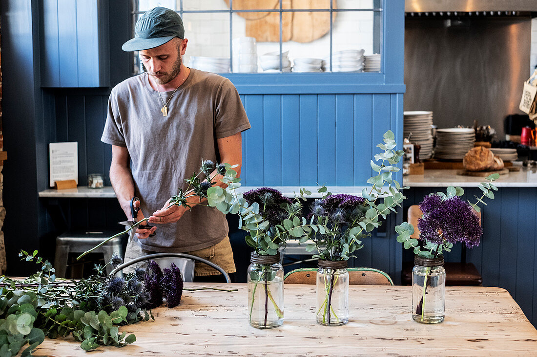 Mann mit Baseballmütze bereitet Blumenarrangement mit Disteln an einem Tisch