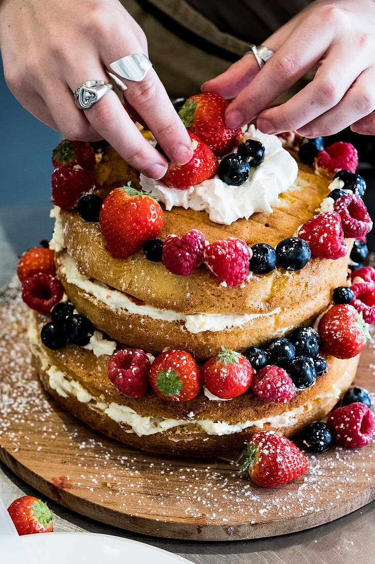 A woman cook asembling a layer cake with fresh cream and fresh fruit, strawberries and blueberries.