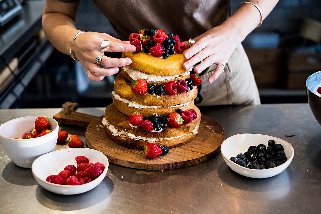 a cook working in a commercial kitchen arranging fresh fruit over a layered cake with fresh cream.