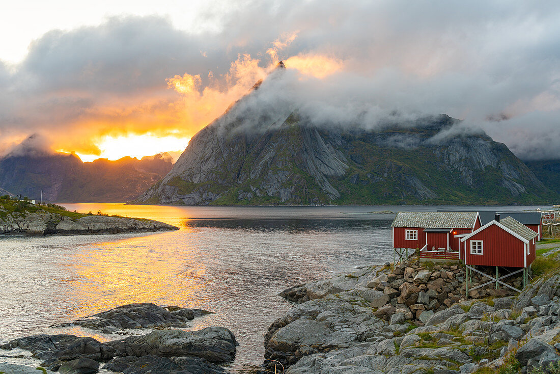 Low clouds swirling round and covering the mountains, a small traditional rorbu house on the shoreline.  Lofoten Islands