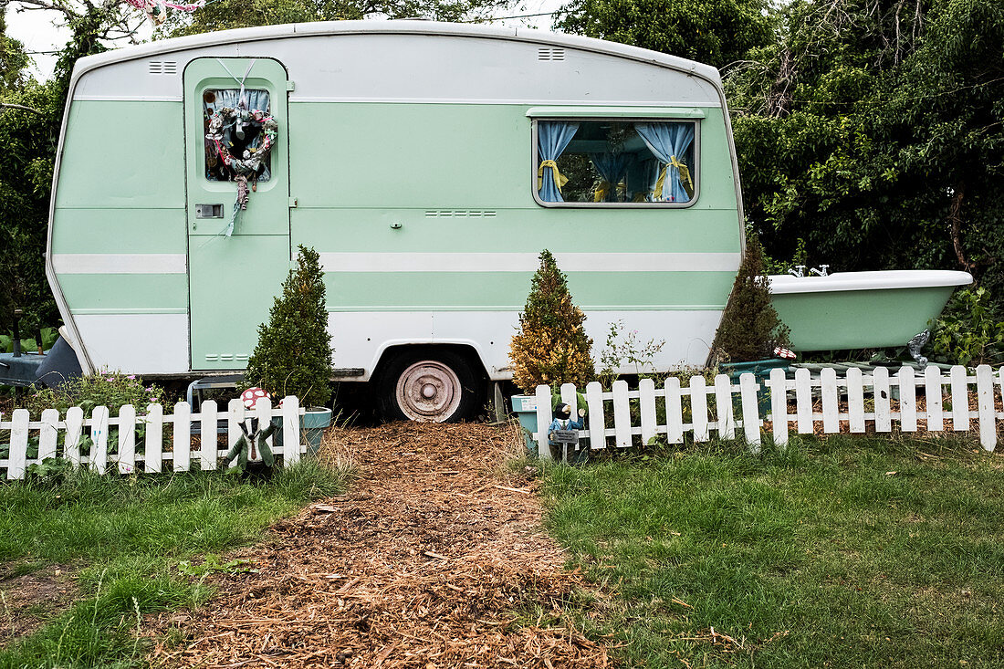 White and green retro caravan parked at the end of a garden path behind low white picket fence.