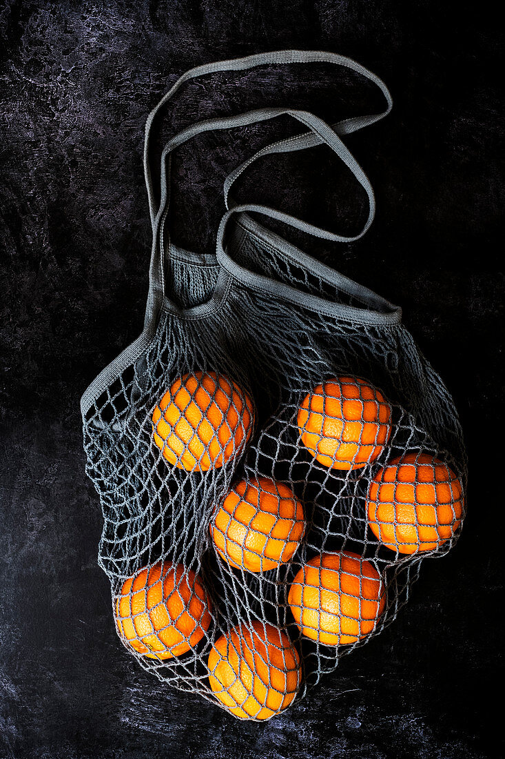 High angle close up of oranges in grey net bag on black background.