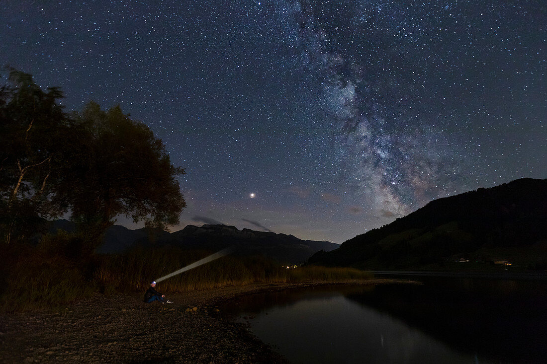 Mann mit Stirnlampe unter Milchstrasse am Nachthimmel über dem Sihlsee, Einsiedeln, Schweiz
