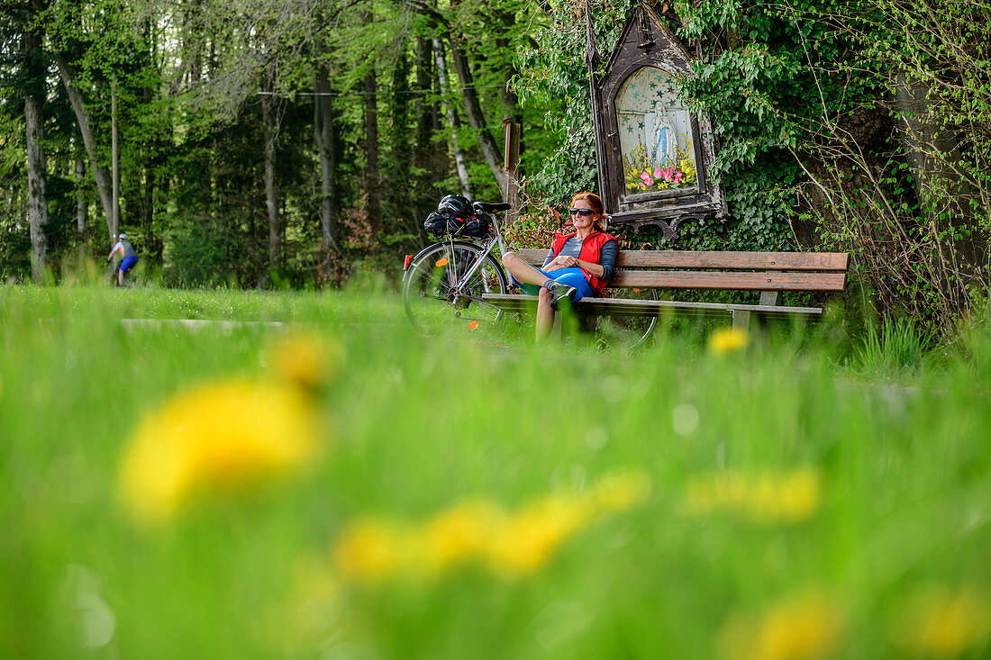Frau beim Radfahren sitzt auf Bank und macht unter Bildstock Pause, Baum-zu-Baum-Radweg, Irschenberg, Oberbayern, Bayern, Deutschland