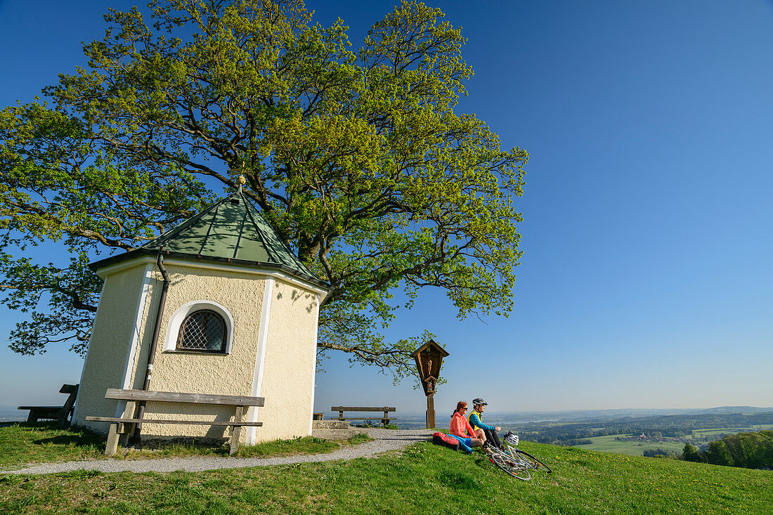 Woman and man cycling while taking break at chapel, Samerberg, Chiemgau, Chiemgau Alps, Upper Bavaria, Bavaria, Germany