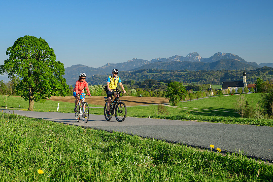 Frau und Mann beim Radfahren, Kirche Wilparting und Mangfallgebirge im Hintergrund, Irschenberg, Oberbayern, Bayern, Deutschland