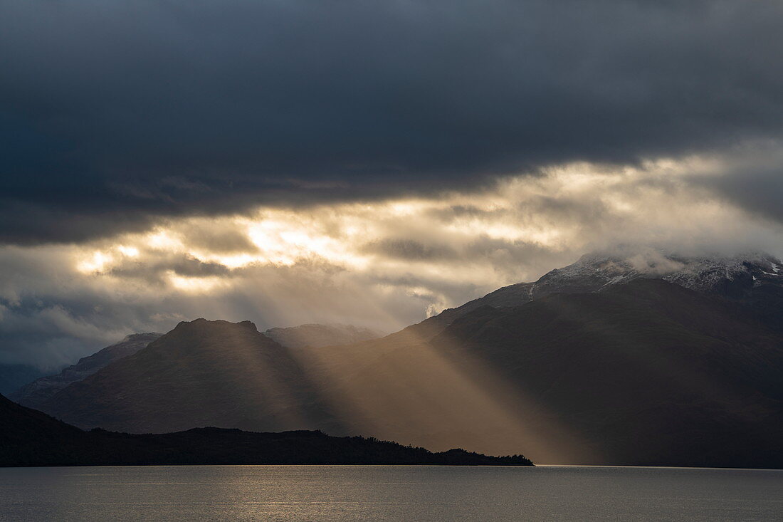 Lichtstrahlen am späten Nachmittag brechen durch eine schwere Wolkendecke über eine Szene aus Bergen und ruhiger See, nahe Pio XI-Gletscher, Magallanes y de la Antartica Chilena, Patagonien, Chile, Südamerika