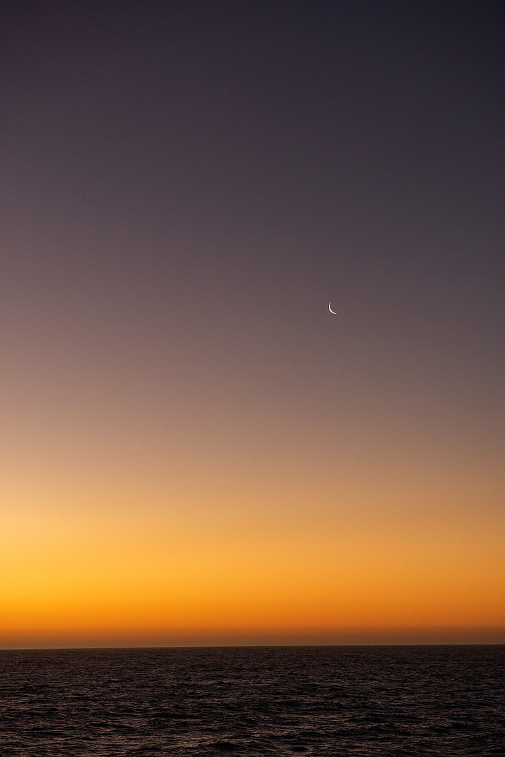 A crescent moon rises as the last warm glow of the sun fades on the horizon, near Puerto Chacabuco, Aysén, Patagonia, Chile, South America