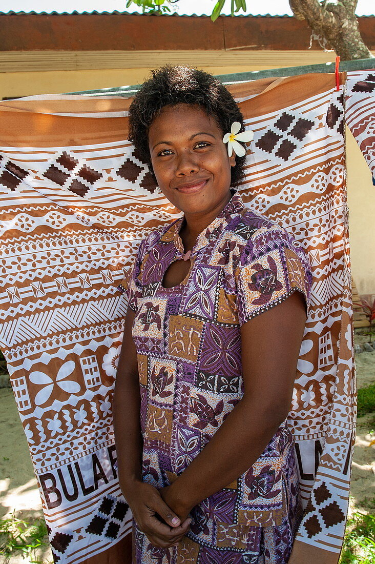 A smiling young woman with a frangipani (plumeria) flower in her hair stands in front of patterned pieces of fabric that she sells to visiting tourists from an expedition cruise ship, Mamanuca Islands, Fiji, South Pacific