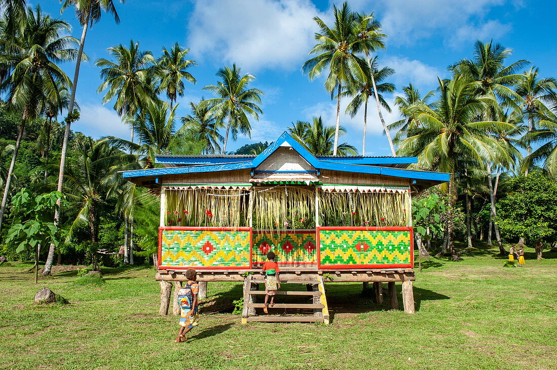 Personen vor einem bunt gemusterten Stelzenhaus  in einer von Palmen umgebenen Rasenlandschaft, Garove Island, Vitu-Inseln, Provinz West New Britain, Papua-Neuguinea, Südpazifik